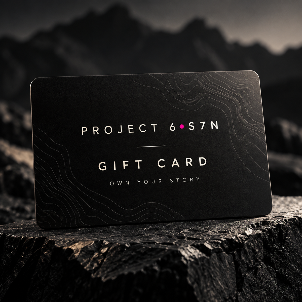 A black gift card with white text reading 'PROJECT 6.S7N GIFT CARD' and 'OWN YOUR STORY' lies on a dark textured surface with blurred mountain scenery in the background.