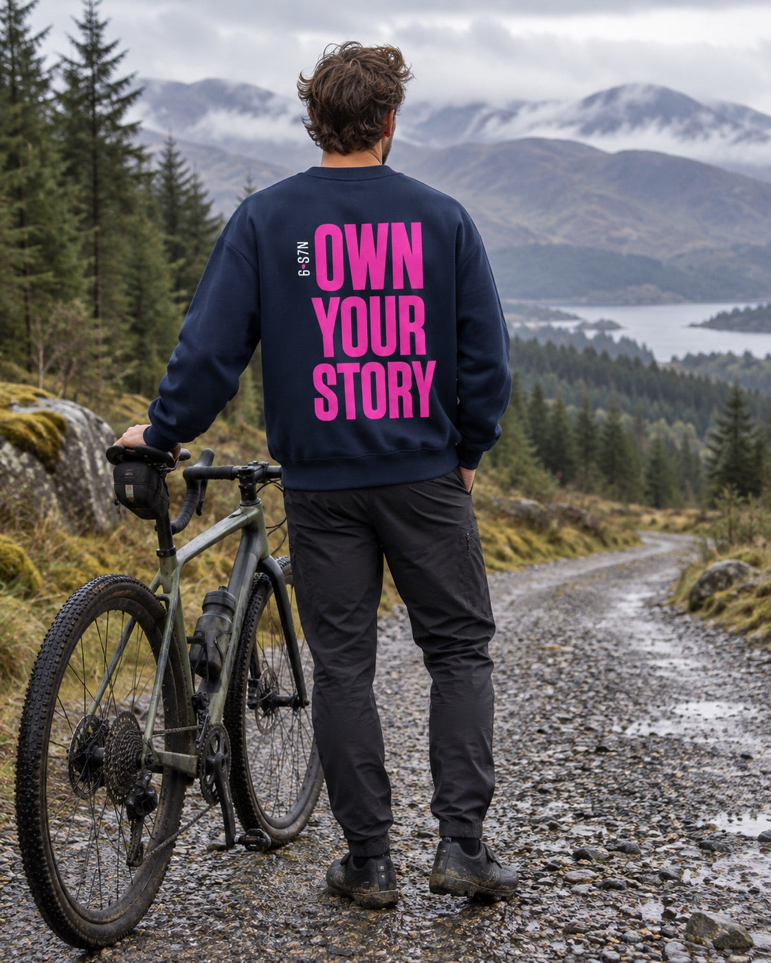 A man stands on a gravel path in a mountain landscape, viewed from behind, wearing a navy sweatshirt with bold pink text that reads 'OWN YOUR STORY' and dark pants, with a bicycle beside him.