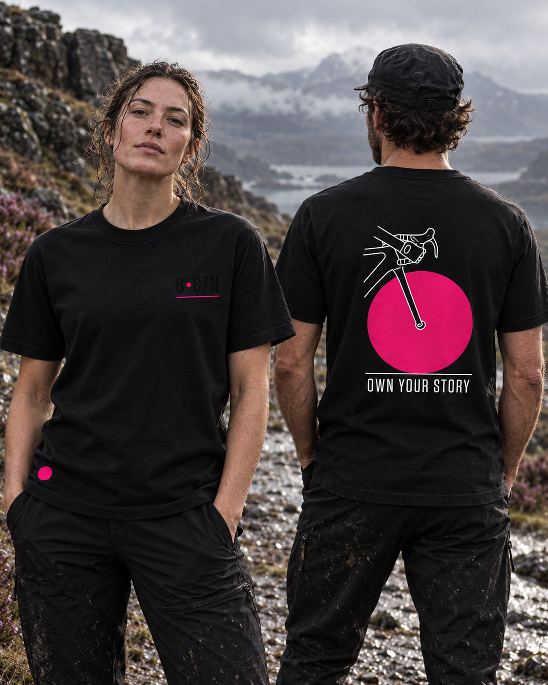 Two models wearing black t-shirts with cycling-themed graphics, standing outdoors in a rocky, mountainous landscape. The woman shows the front of the shirt with a small chest logo, and the man shows the back with a large pink circle, bicycle graphic, and the text 'OWN YOUR STORY'.